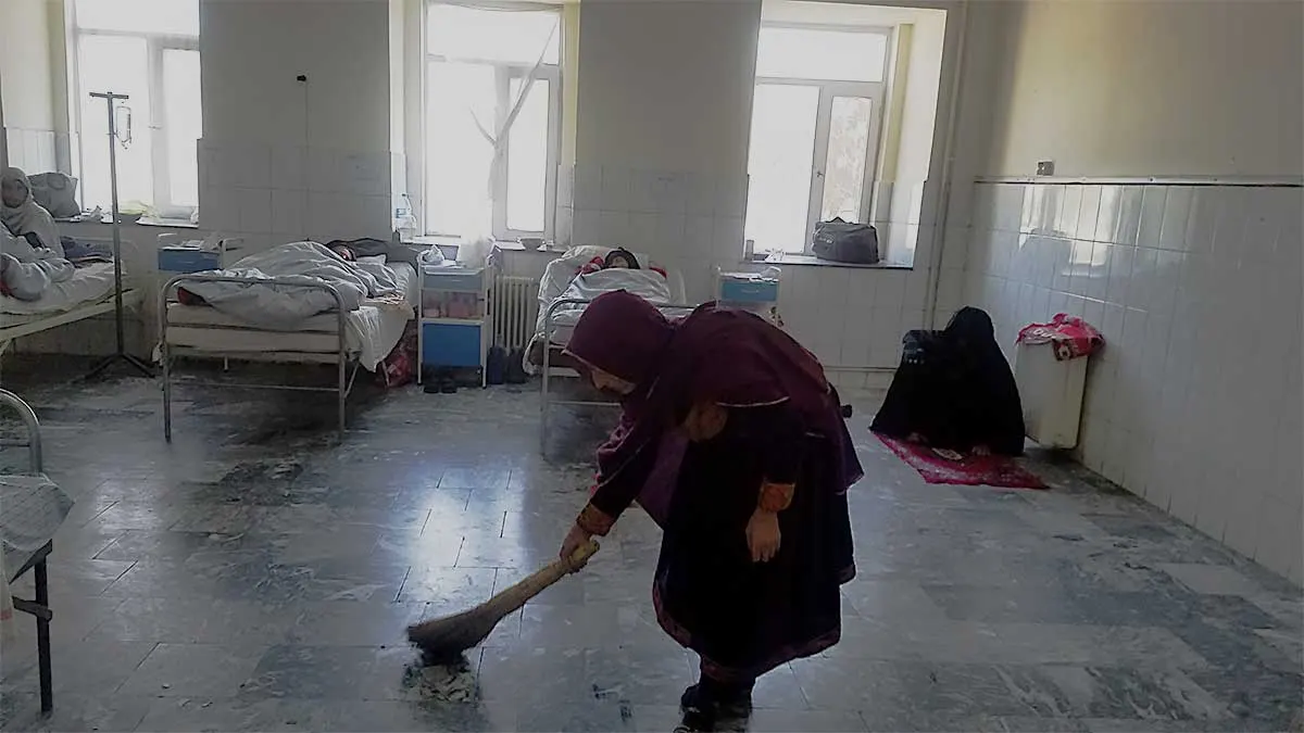 Old woman cleaning hospital ward with patients in bed
